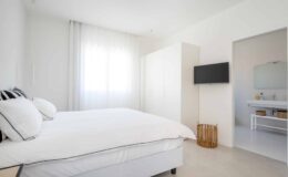 Bright and modern bedroom featuring a queen-sized bed with white linens, a flat-screen TV mounted on the wall, and a spacious wardrobe. Natural light filters through sheer curtains, creating a serene atmosphere. A glimpse of a stylish bathroom is visible through an open doorway.