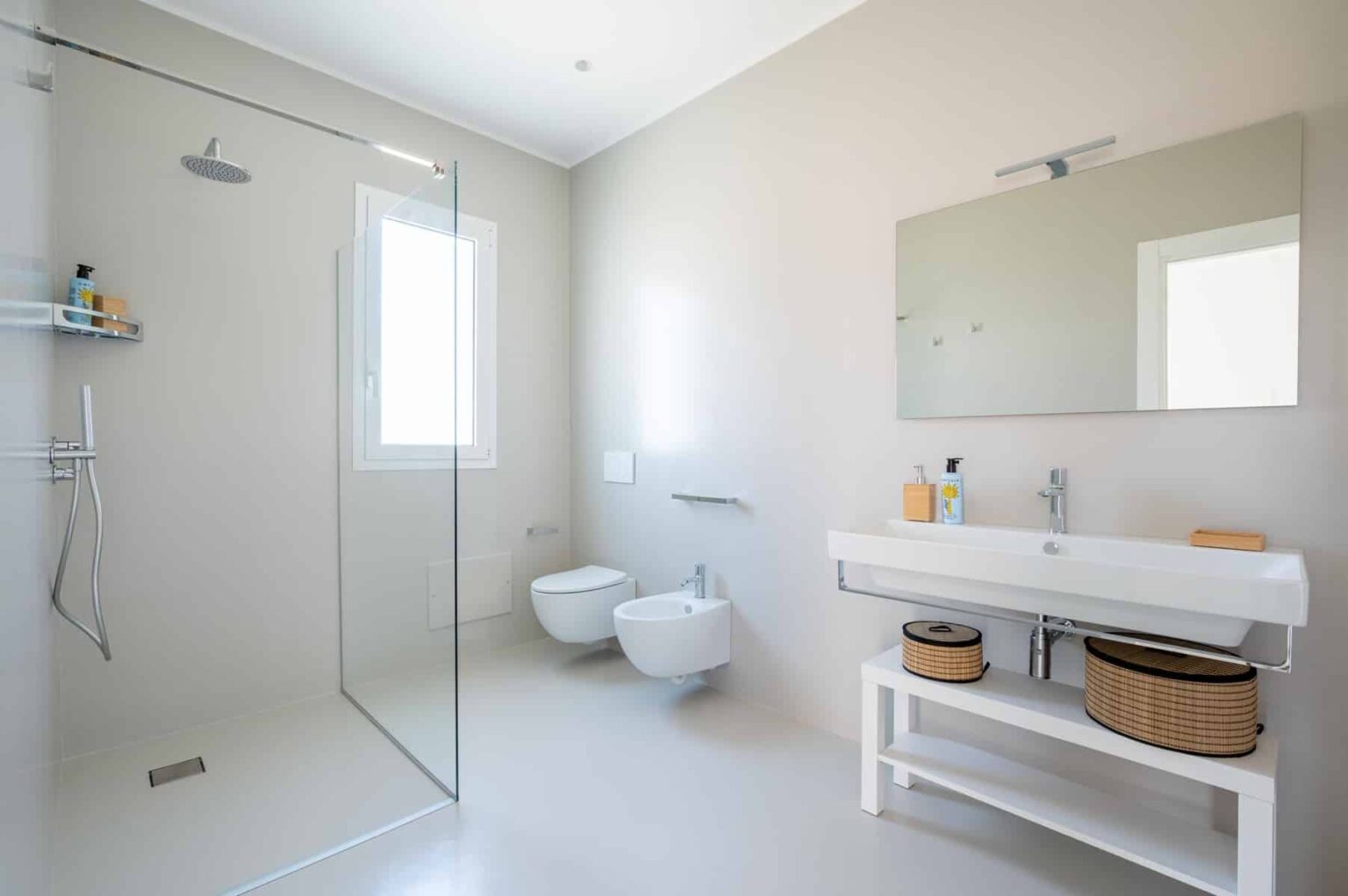 Modern bathroom featuring a glass shower, minimalist design, a wall-mounted sink with a wooden storage basket, and a large mirror, all set against neutral-colored walls. | Only on SopranoVillas