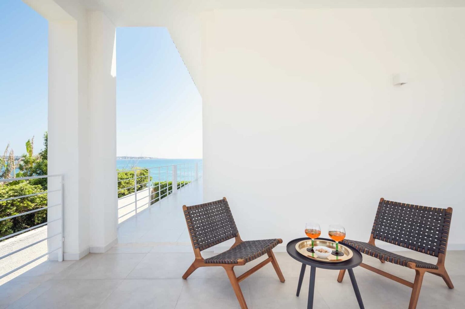 Modern balcony with two wooden chairs and a small table, featuring drinks and snacks, overlooking a scenic ocean view. Bright, minimalist design with white walls and natural light. | Only on SopranoVillas