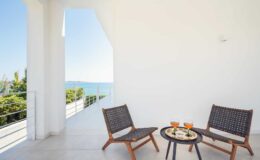 Modern balcony with two wooden chairs and a small table, featuring drinks and snacks, overlooking a scenic ocean view. Bright, minimalist design with white walls and natural light.