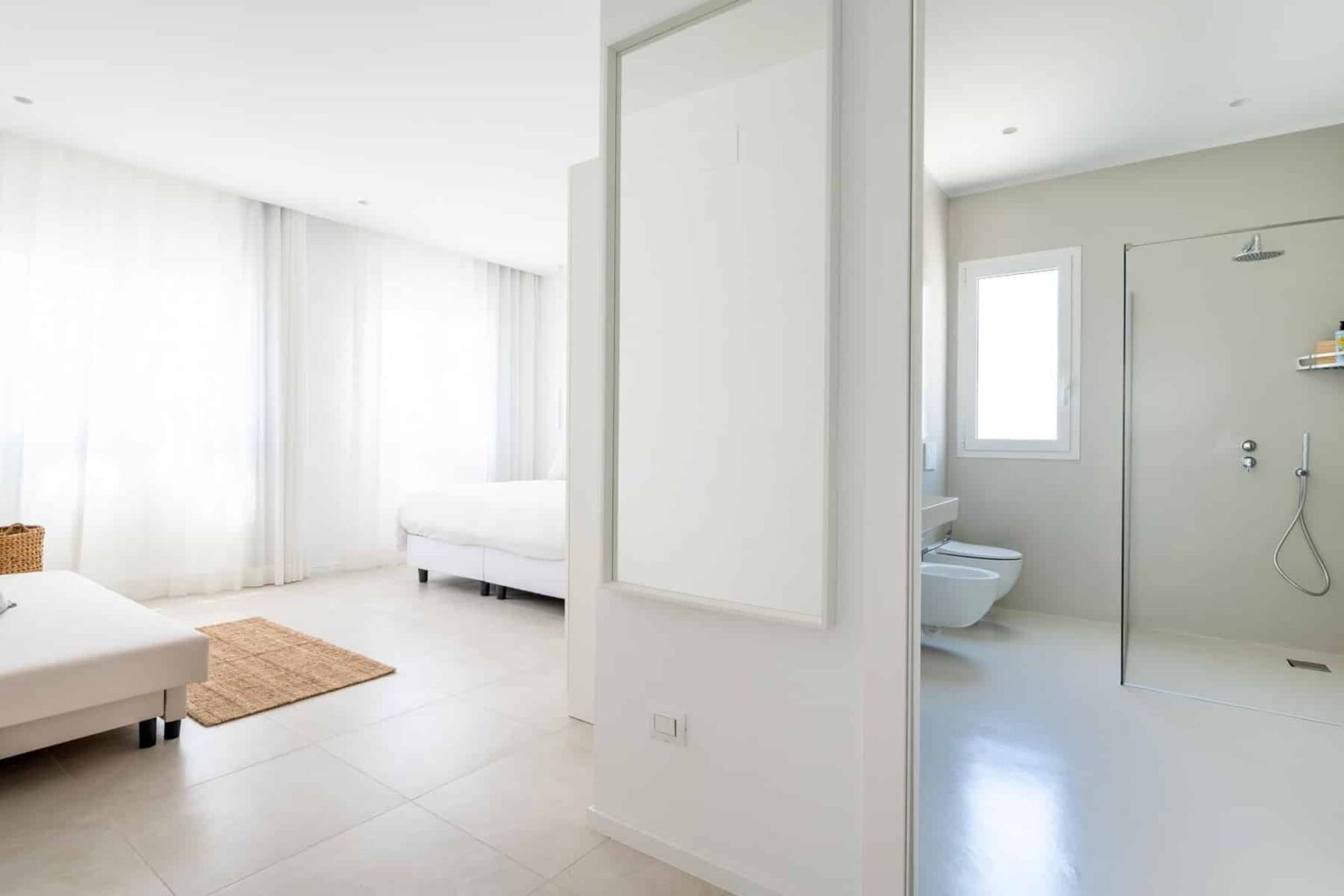 Bright and modern bedroom with two beds, large windows allowing natural light, and an adjoining bathroom featuring a shower. The minimalist design includes neutral colors and clean lines, creating a spacious and inviting atmosphere. | Only on SopranoVillas