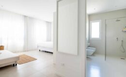 Bright and modern bedroom with two beds, large windows allowing natural light, and an adjoining bathroom featuring a shower. The minimalist design includes neutral colors and clean lines, creating a spacious and inviting atmosphere.