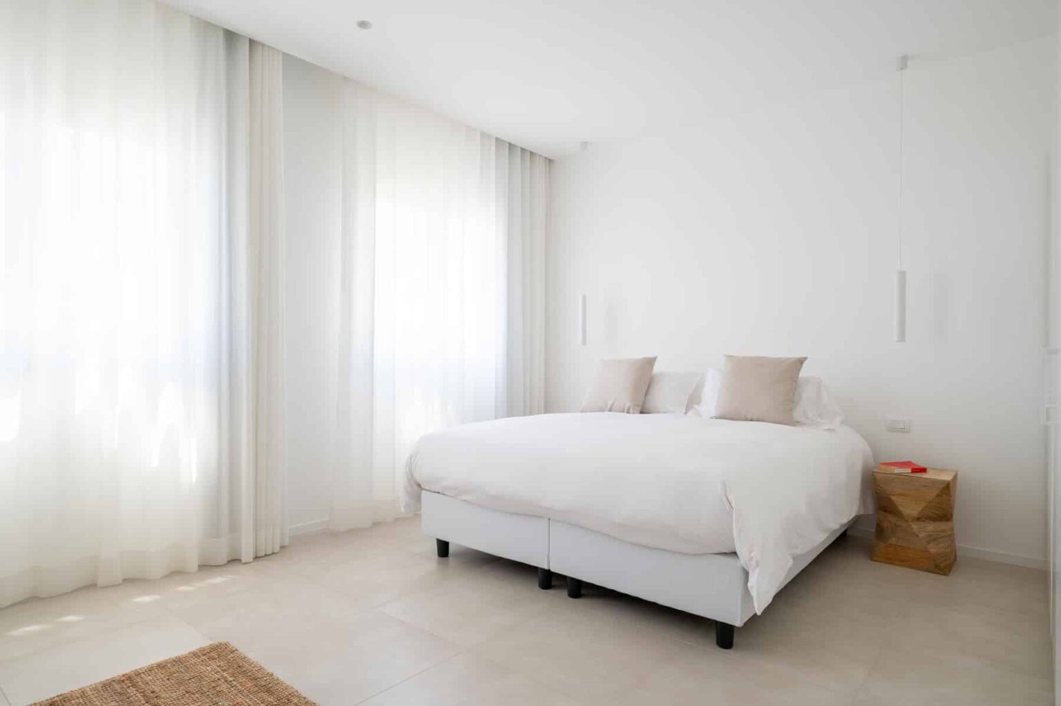 Bright and airy modern bedroom featuring a large white bed with two beige pillows, light sheer curtains, and a wooden nightstand, creating a serene and minimalist atmosphere. | Only on SopranoVillas