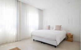 Bright and airy modern bedroom featuring a large white bed with two beige pillows, light sheer curtains, and a wooden nightstand, creating a serene and minimalist atmosphere.