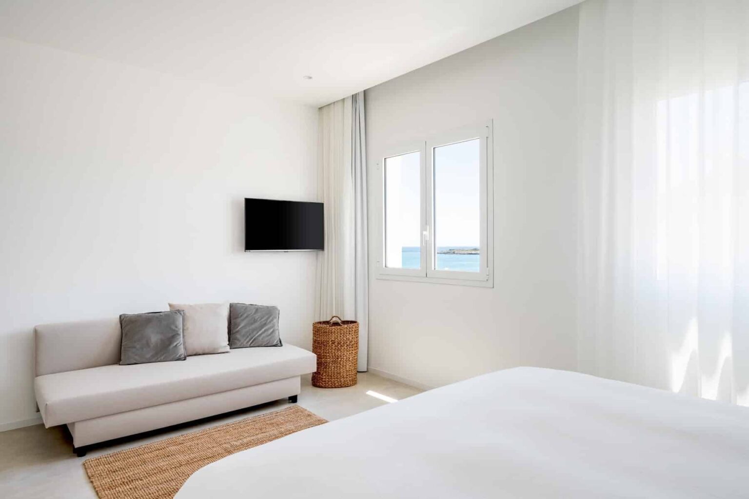 Modern bedroom interior featuring a light color palette, a cozy sofa with decorative pillows, a stylish wicker basket, and a wall-mounted TV. Large windows allow for natural light, enhancing the airy atmosphere. | Only on SopranoVillas
