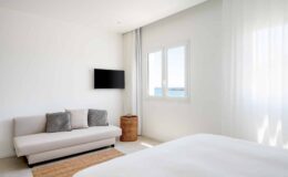 Modern bedroom interior featuring a light color palette, a cozy sofa with decorative pillows, a stylish wicker basket, and a wall-mounted TV. Large windows allow for natural light, enhancing the airy atmosphere.