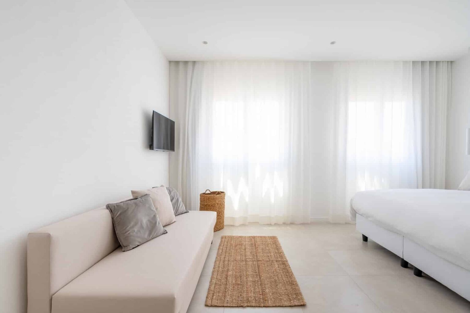 Modern minimalist bedroom featuring a light-colored sofa with cushions, a wall-mounted TV, and large windows with sheer curtains allowing natural light, complemented by a woven area rug. | Only on SopranoVillas