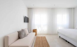 Modern minimalist bedroom featuring a light-colored sofa with cushions, a wall-mounted TV, and large windows with sheer curtains allowing natural light, complemented by a woven area rug.