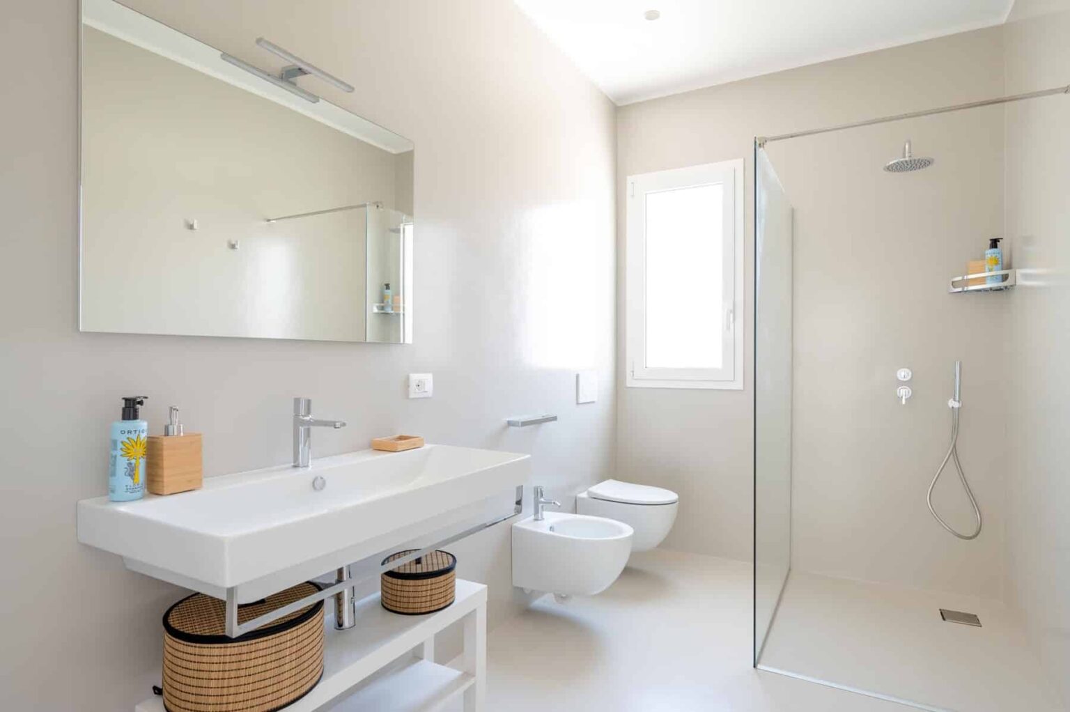Modern bathroom with a sleek design featuring a glass shower, minimalist white vanity, and natural light from a window. The space includes a wall-mounted mirror, stylish toiletries, and decorative storage baskets. | Only on SopranoVillas