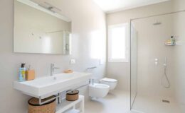 Modern bathroom with a sleek design featuring a glass shower, minimalist white vanity, and natural light from a window. The space includes a wall-mounted mirror, stylish toiletries, and decorative storage baskets.