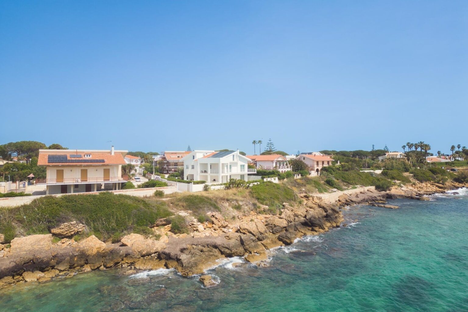 Coastal view of residential homes along a rocky shoreline with clear turquoise waters and a bright blue sky, highlighting a serene seaside environment. | Only on SopranoVillas