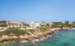 Coastal view of residential homes along a rocky shoreline with clear turquoise waters and a bright blue sky, highlighting a serene seaside environment.