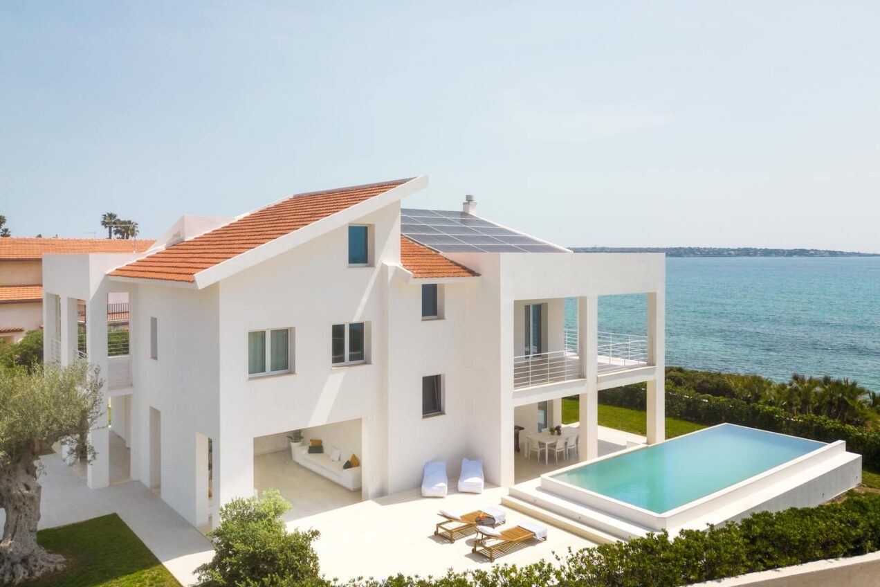 Modern white beachfront villa with a solar panel roof, featuring a swimming pool, outdoor seating area, and lush greenery, overlooking the ocean. | Only on SopranoVillas