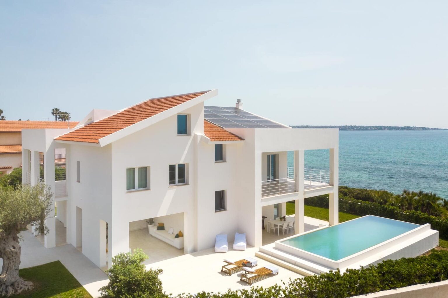 Modern white beachfront villa with a solar panel roof, featuring a swimming pool, outdoor seating area, and lush greenery, overlooking the ocean. | Only on SopranoVillas