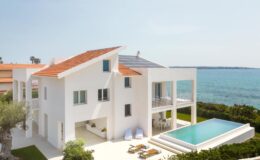 Modern white beachfront villa with a solar panel roof, featuring a swimming pool, outdoor seating area, and lush greenery, overlooking the ocean.