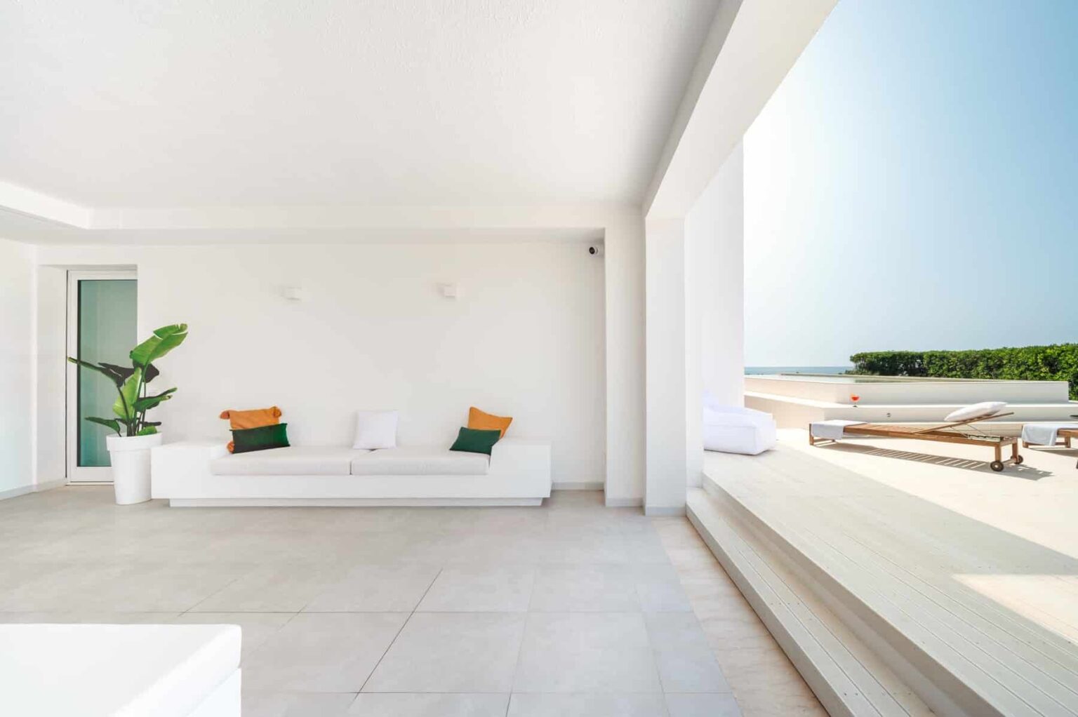 Modern minimalist living room with a white sofa, green plant, and large windows offering a view of the outdoors. The space features neutral tones and natural light, creating a serene atmosphere. | Only on SopranoVillas
