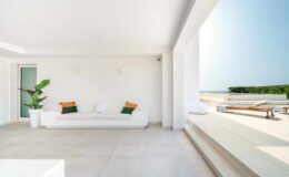 Modern minimalist living room with a white sofa, green plant, and large windows offering a view of the outdoors. The space features neutral tones and natural light, creating a serene atmosphere.