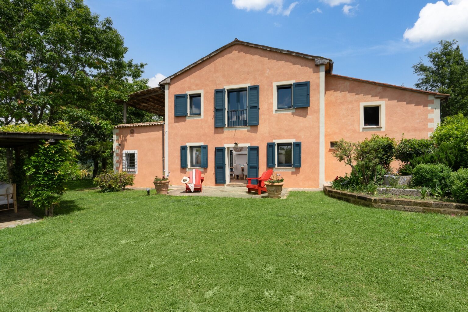 Charming two-story brick house with blue shutters surrounded by a lush green lawn and landscaped garden, featuring outdoor seating areas and trees under a clear blue sky. | Only on SopranoVillas