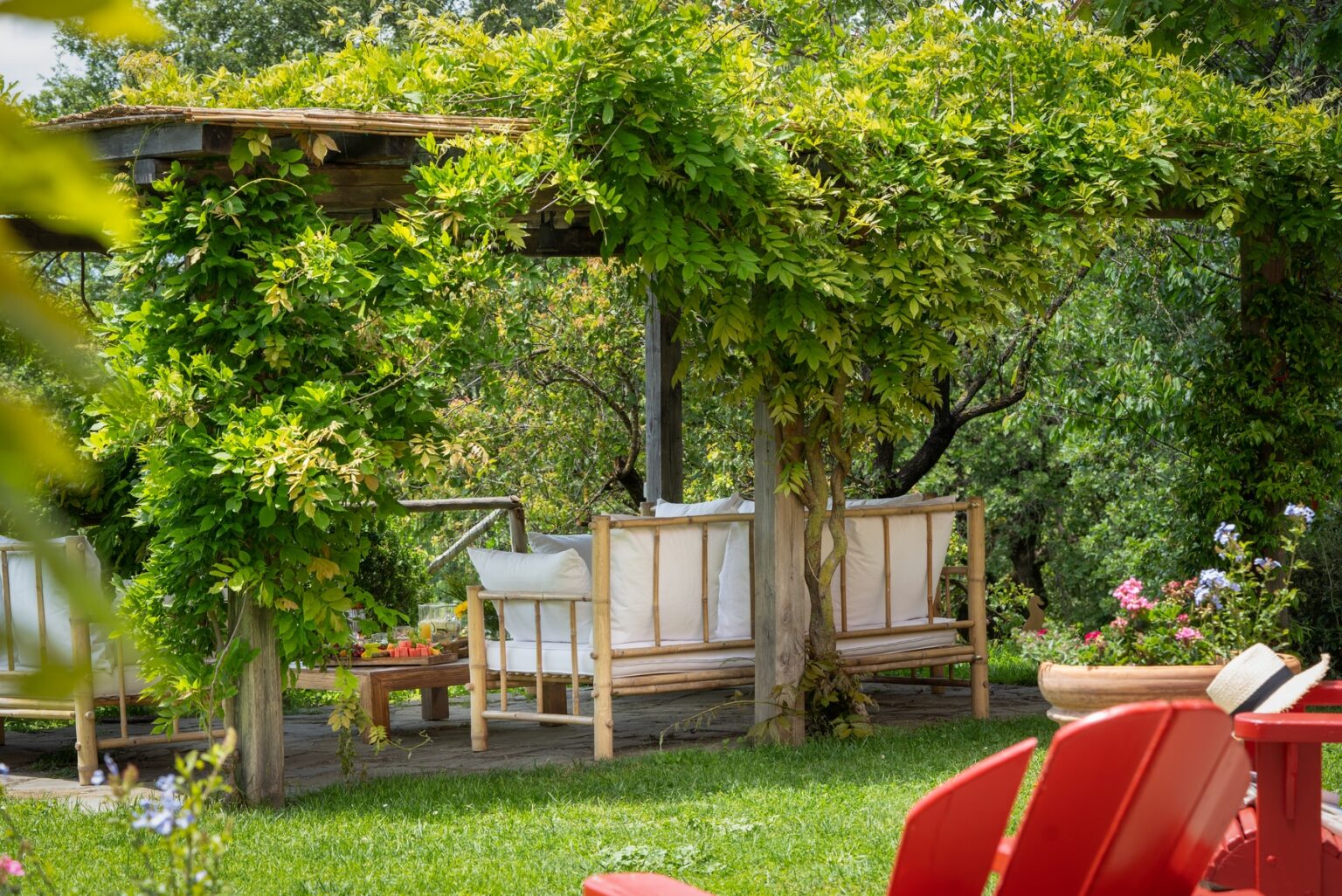 Cozy outdoor seating area under a lush green pergola, featuring a wooden bench with white cushions, a small table, and vibrant flowers in the background, creating a serene garden retreat. | Only on SopranoVillas