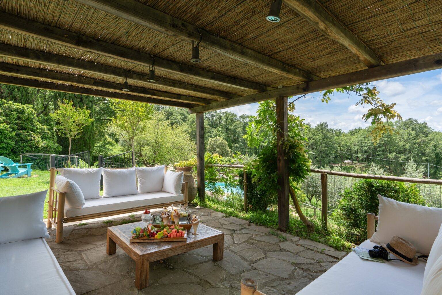 Cozy outdoor seating area with white sofas and a wooden coffee table, surrounded by lush greenery and a scenic view of nature under a thatched roof. Perfect for relaxation and entertaining guests. | Only on SopranoVillas