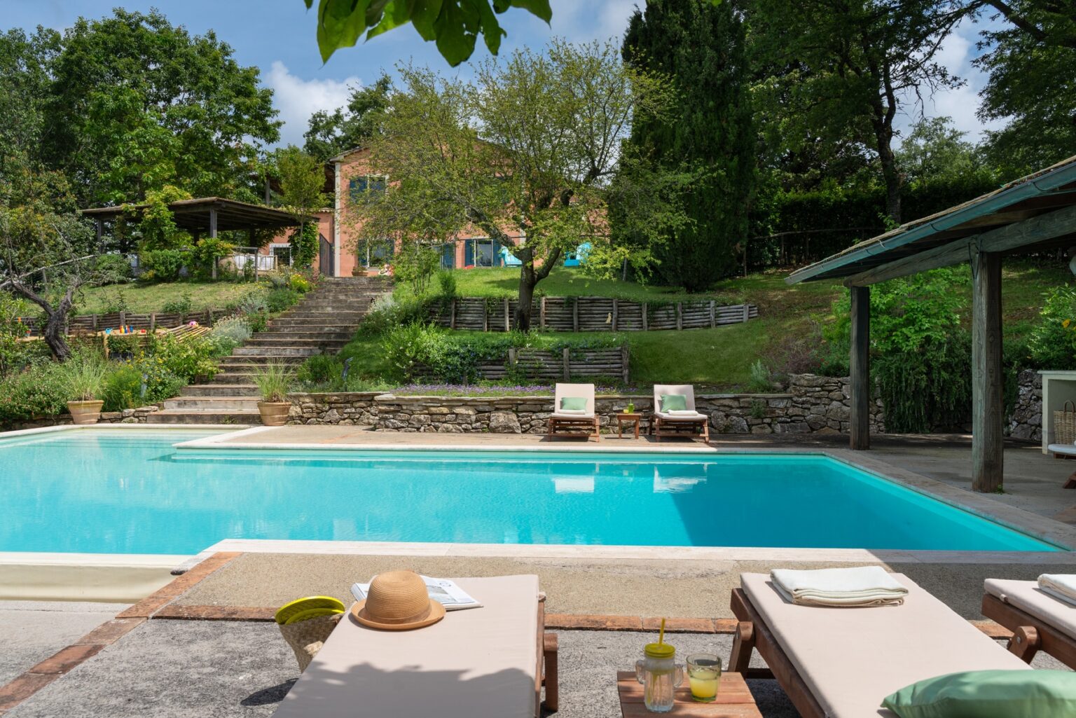 Luxurious outdoor pool area surrounded by lush greenery, featuring comfortable lounge chairs and a charming house in the background, perfect for relaxation and leisure. | Only on SopranoVillas