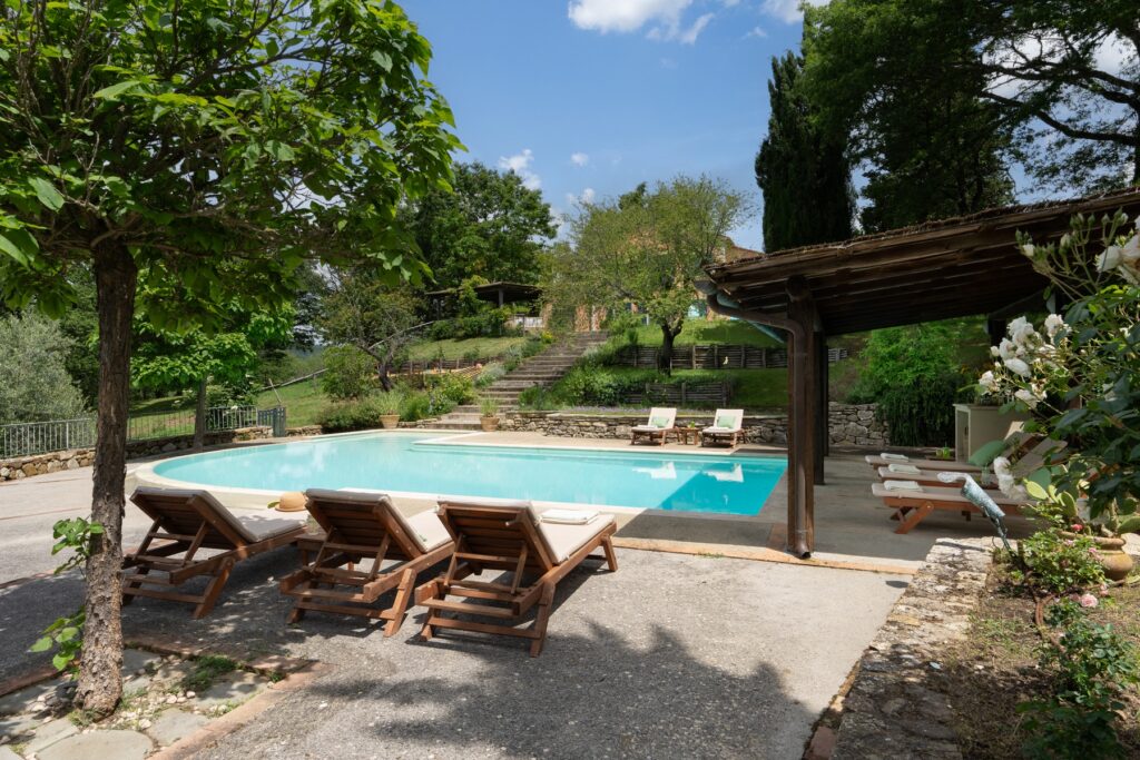 Serene outdoor swimming pool area surrounded by lush greenery, featuring comfortable lounge chairs and a rustic wooden pergola, perfect for relaxation in a tranquil setting. | Only on SopranoVillas