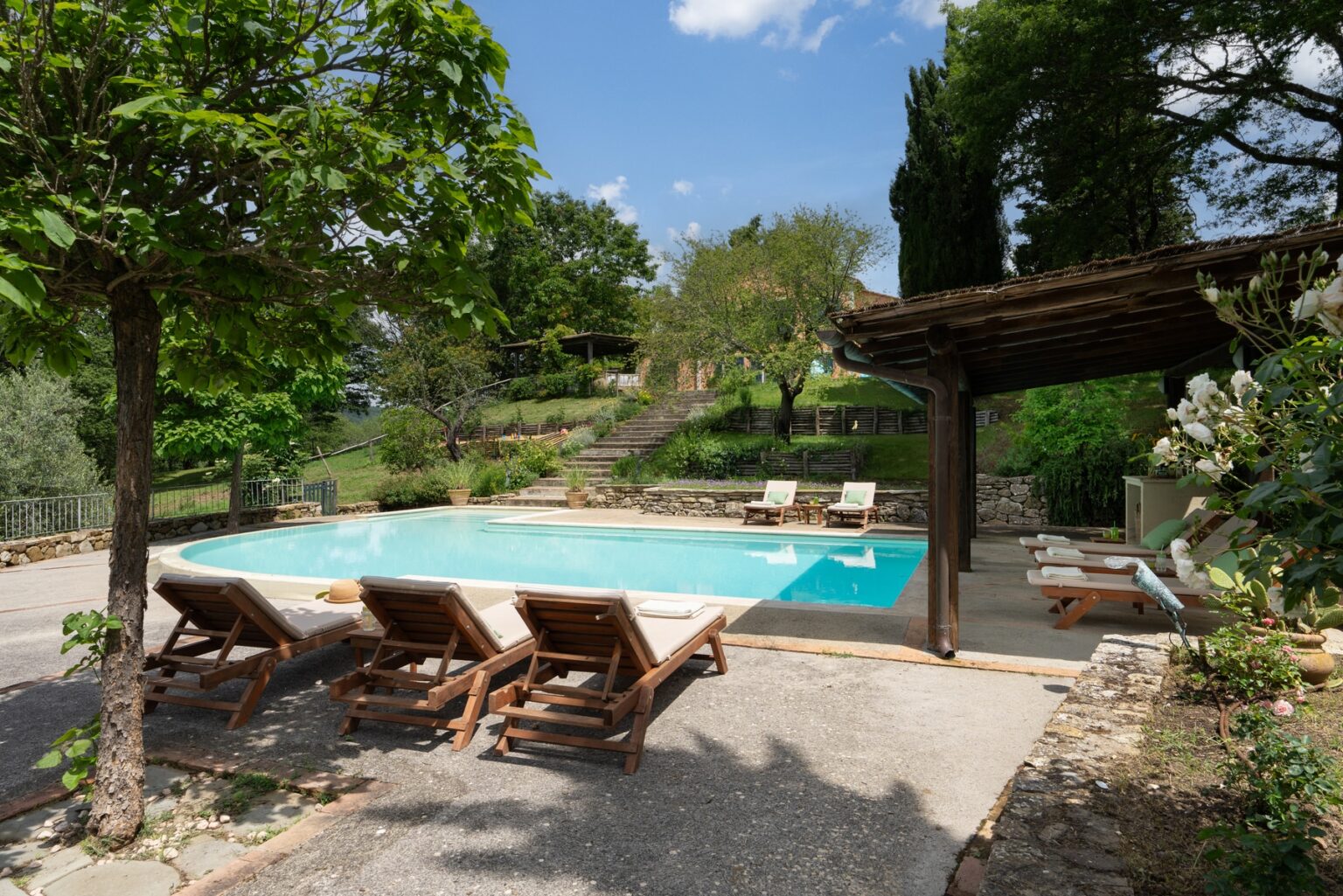 Serene outdoor swimming pool area surrounded by lush greenery, featuring comfortable lounge chairs and a rustic wooden pergola, perfect for relaxation in a tranquil setting. | Only on SopranoVillas