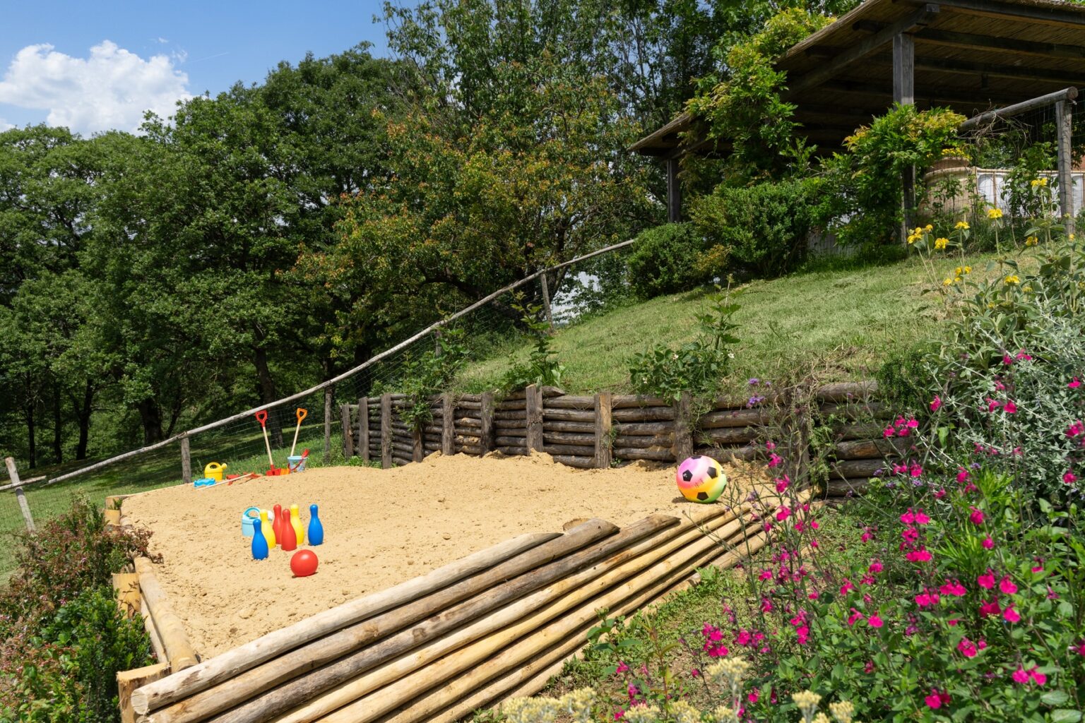 Children's sand play area featuring colorful toys, including shovels, bowling pins, and a ball, surrounded by lush greenery and flowers. Ideal for outdoor activities and playtime in a natural setting. | Only on SopranoVillas