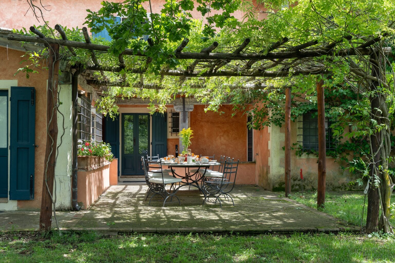 Outdoor dining area under a lush pergola with green vines, featuring a round table set for a meal, surrounded by chairs, against a backdrop of a rustic home with warm-colored walls and blue shutters. Ideal for gatherings and enjoying nature. | Only on SopranoVillas