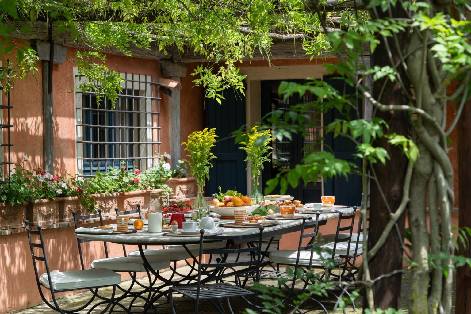 Outdoor dining setup with a beautifully arranged table featuring food and drinks, surrounded by greenery and a cozy, rustic house. Perfect for a summer gathering or brunch. | Only on SopranoVillas