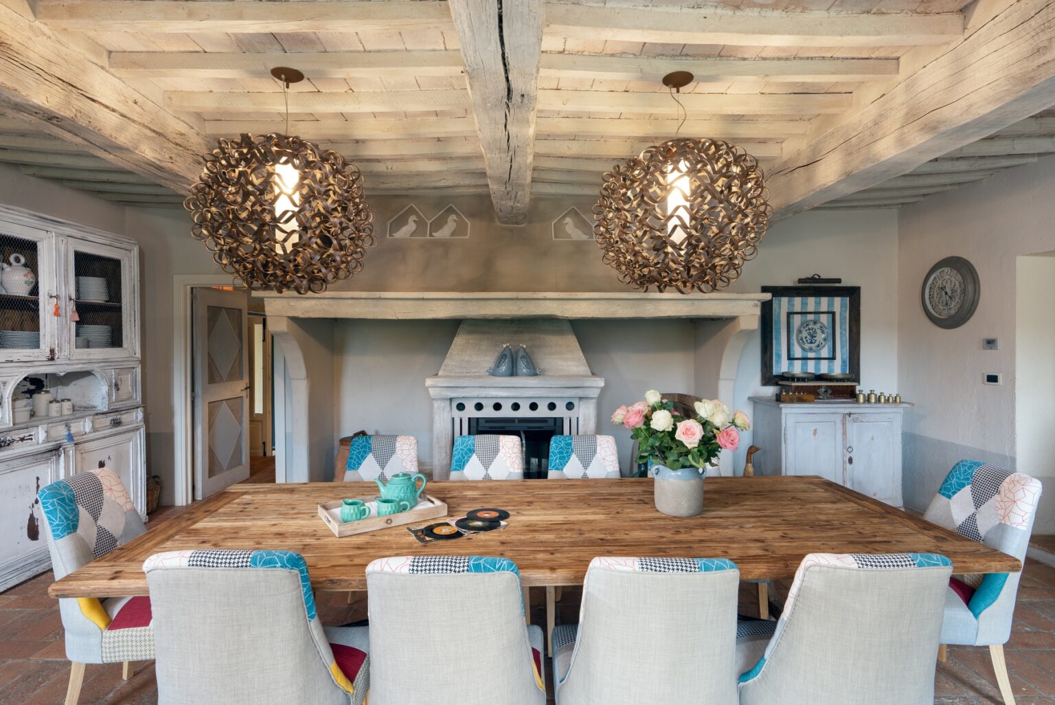 Spacious dining room featuring a large wooden table surrounded by upholstered chairs, decorative floral centerpiece, and stylish pendant lighting. The room has a rustic charm with exposed wooden beams and a cozy fireplace, creating an inviting atmosphere for gatherings. | Only on SopranoVillas