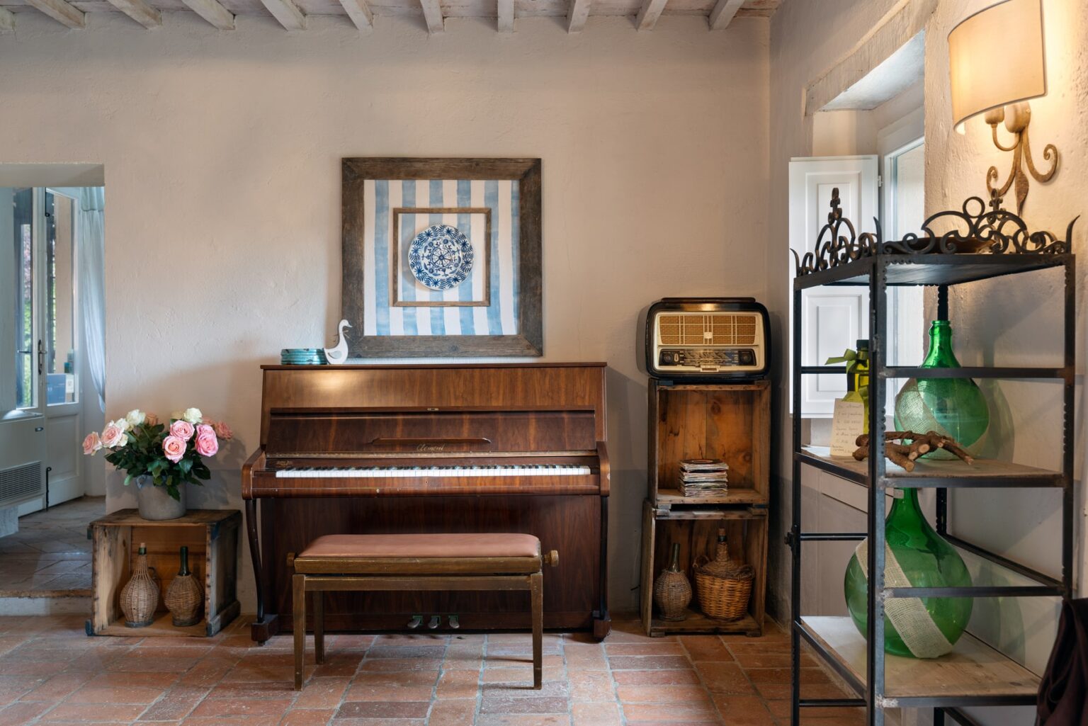 Cozy living room featuring a vintage piano with a wooden bench, floral arrangement, and retro radio. Decorated with blue and white wall art, a metal shelving unit displaying green glass bottles, and rustic wooden accents. Natural light streams in through a window, enhancing the inviting atmosphere. | Only on SopranoVillas