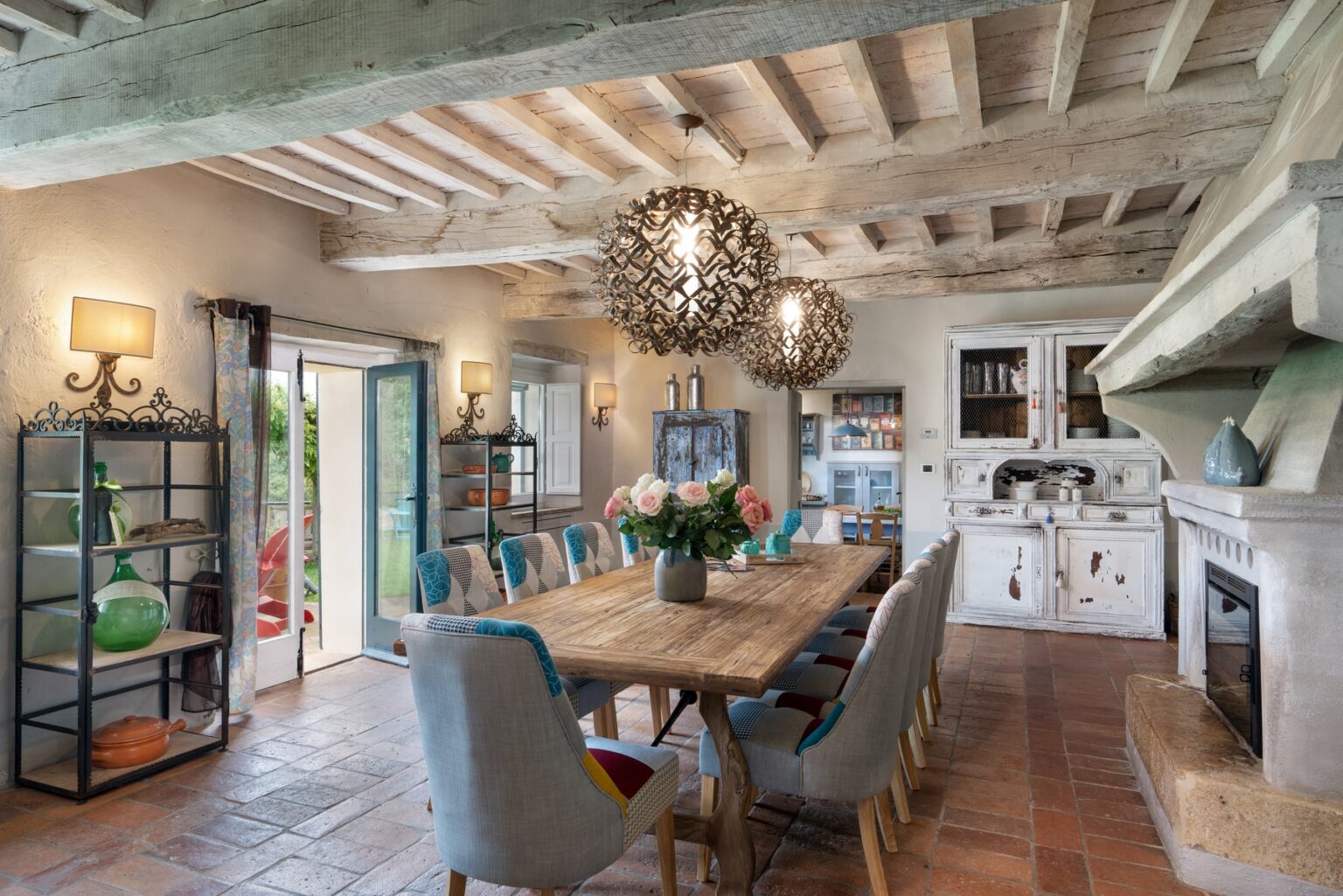 Spacious dining room featuring a rustic wooden table surrounded by upholstered chairs, stylish pendant lighting, and a cozy fireplace. The room has exposed wooden beams and large windows that provide natural light, creating a warm and inviting atmosphere. | Only on SopranoVillas