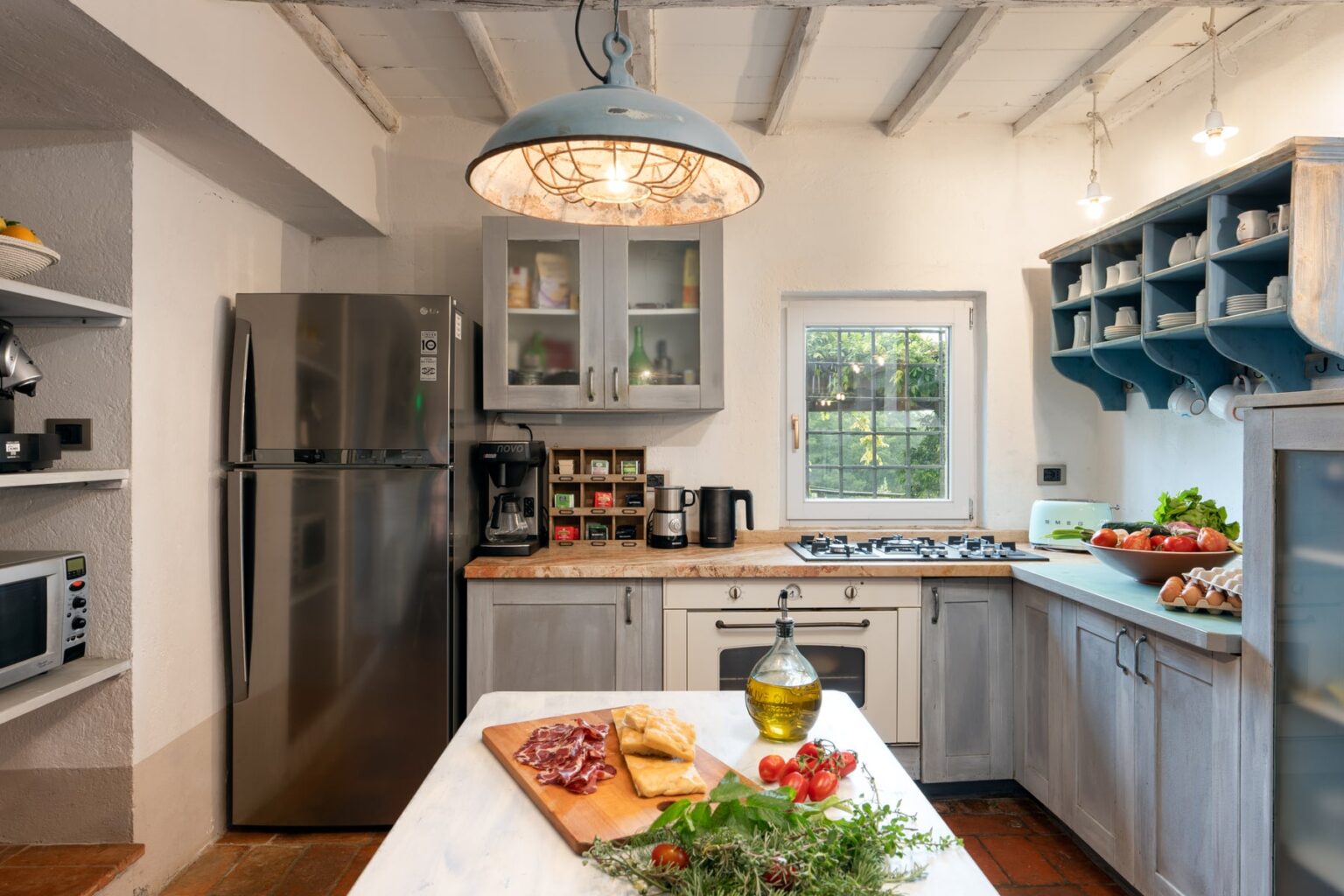 Modern kitchen interior featuring stainless steel appliances, a gas stove, and wooden countertops. Fresh ingredients like herbs, tomatoes, and olive oil are displayed on a cutting board, with open shelves showcasing kitchenware. Natural light floods the space through a window, enhancing the inviting atmosphere. | Only on SopranoVillas