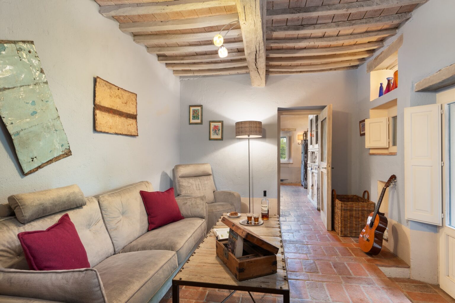 Cozy living room featuring a comfortable gray sofa with red cushions, a wooden coffee table with snacks and drinks, and rustic decor elements. The room has exposed wooden beams, terracotta flooring, and a warm ambiance created by a stylish lamp. Visible in the background are an open kitchen area and a guitar leaning against the wall. | Only on SopranoVillas