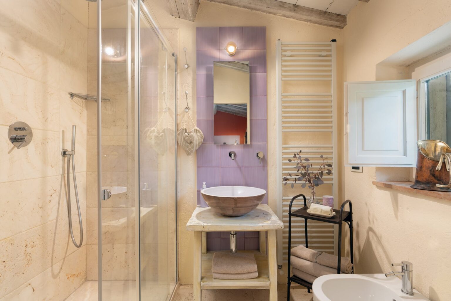 Modern bathroom featuring a glass shower, elegant sink on a wooden stand, and stylish decor with light beige and purple accents. Natural light is provided by a window, enhancing the inviting atmosphere. | Only on SopranoVillas
