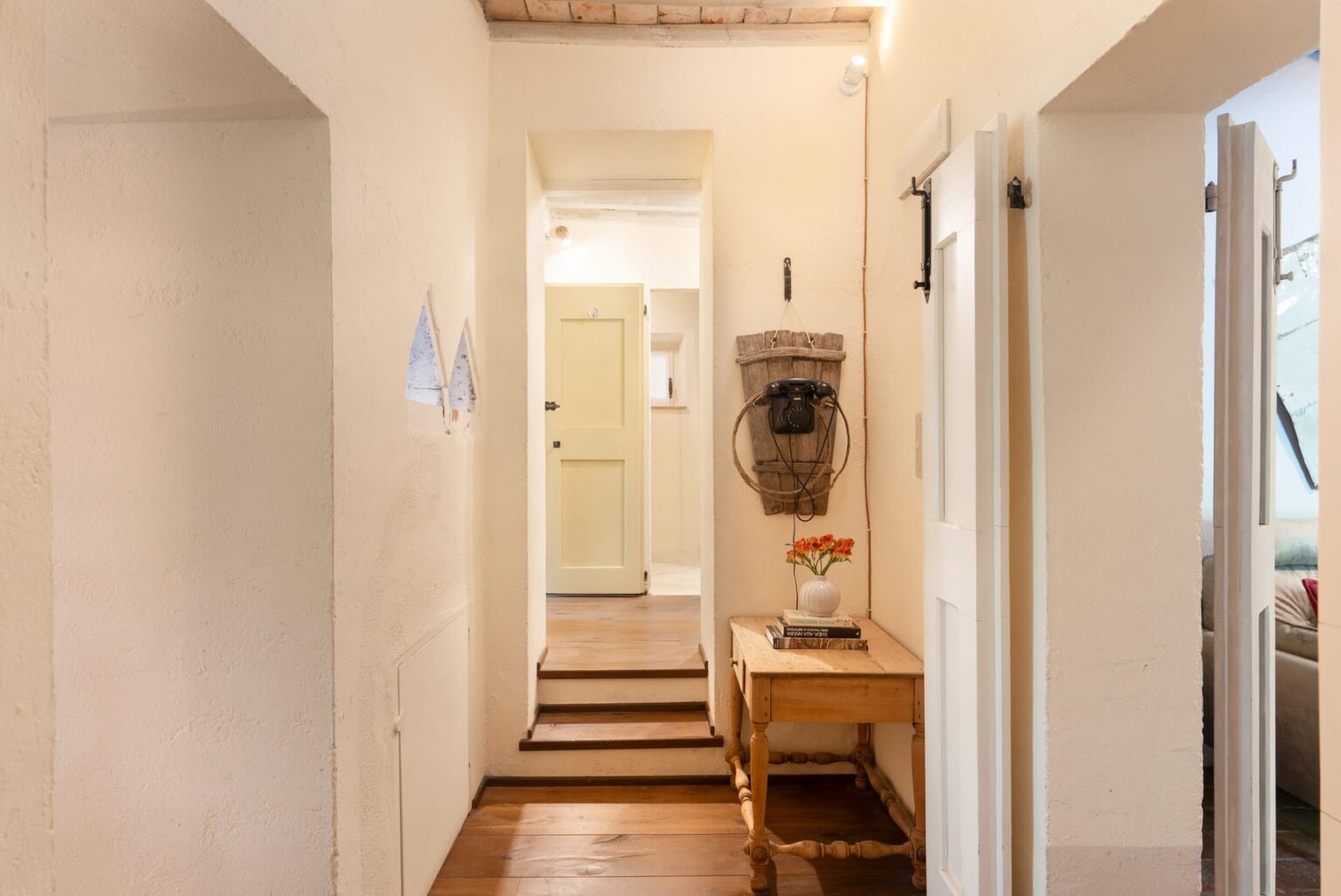 Cozy hallway featuring light-colored walls, wooden flooring, and a rustic table with a vase of flowers. A vintage telephone hangs on the wall beside a yellow door, leading to another room. The space is illuminated by soft lighting, creating a warm and inviting atmosphere. | Only on SopranoVillas