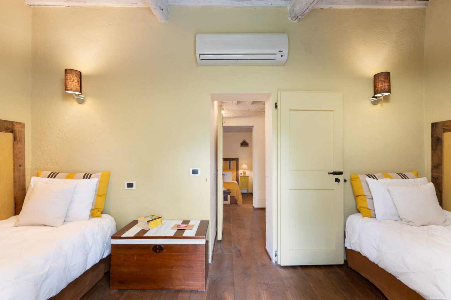 Cozy guest room featuring two single beds with yellow and white bedding, a wooden chest as a nightstand, and warm wall sconces. The room has a light green color scheme and an air conditioning unit, with a doorway leading to a second bedroom. Ideal for travelers seeking comfort and style. | Only on SopranoVillas