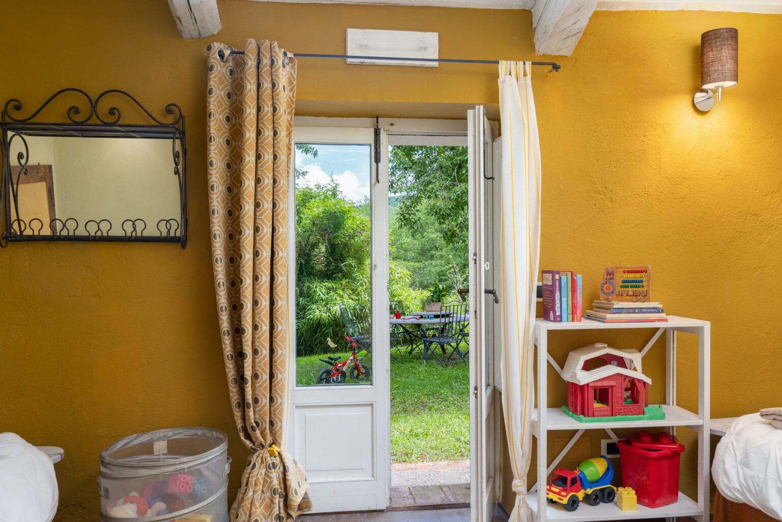 Cozy interior of a brightly colored room featuring a yellow wall, open doors leading to a lush garden, a decorative mirror, and organized shelves with toys and books. | Only on SopranoVillas
