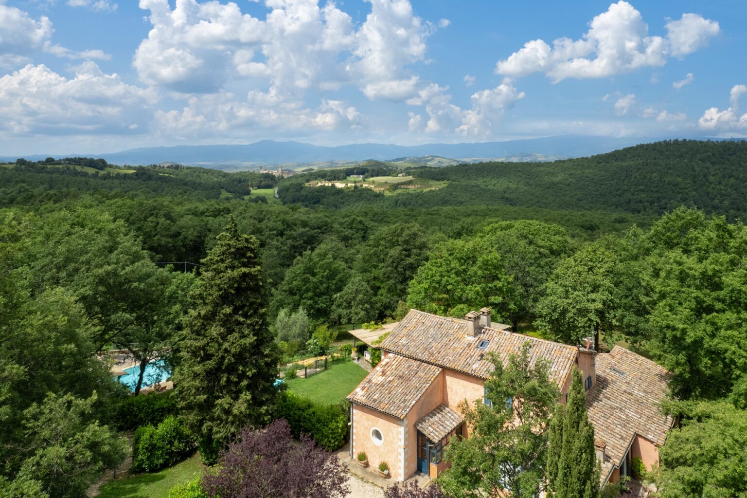 Scenic view of a rustic villa surrounded by lush green forests and rolling hills under a partly cloudy sky, showcasing a tranquil countryside landscape. | Only on SopranoVillas