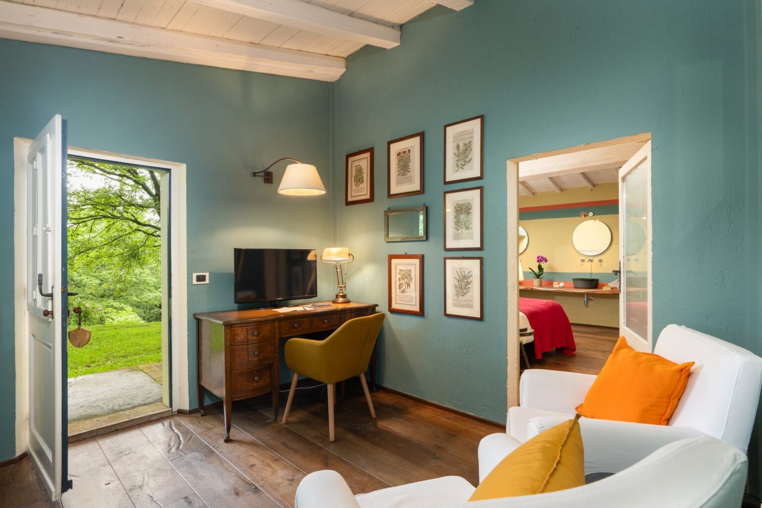 Cozy interior of a modern living space with teal walls, featuring a wooden desk, a comfortable yellow chair, and white sofas with orange accents. A door opens to a lush outdoor view, while framed artwork adorns the walls, enhancing the inviting atmosphere. | Only on SopranoVillas