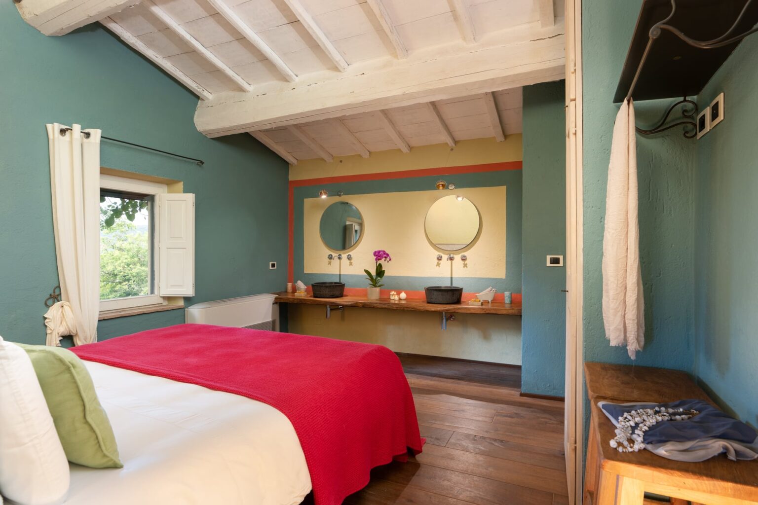Brightly colored bedroom featuring a red bedspread, wooden flooring, and large mirrors above a stylish vanity. The walls are painted in calming blue tones, complemented by natural light from a window, creating a welcoming and relaxing atmosphere. | Only on SopranoVillas