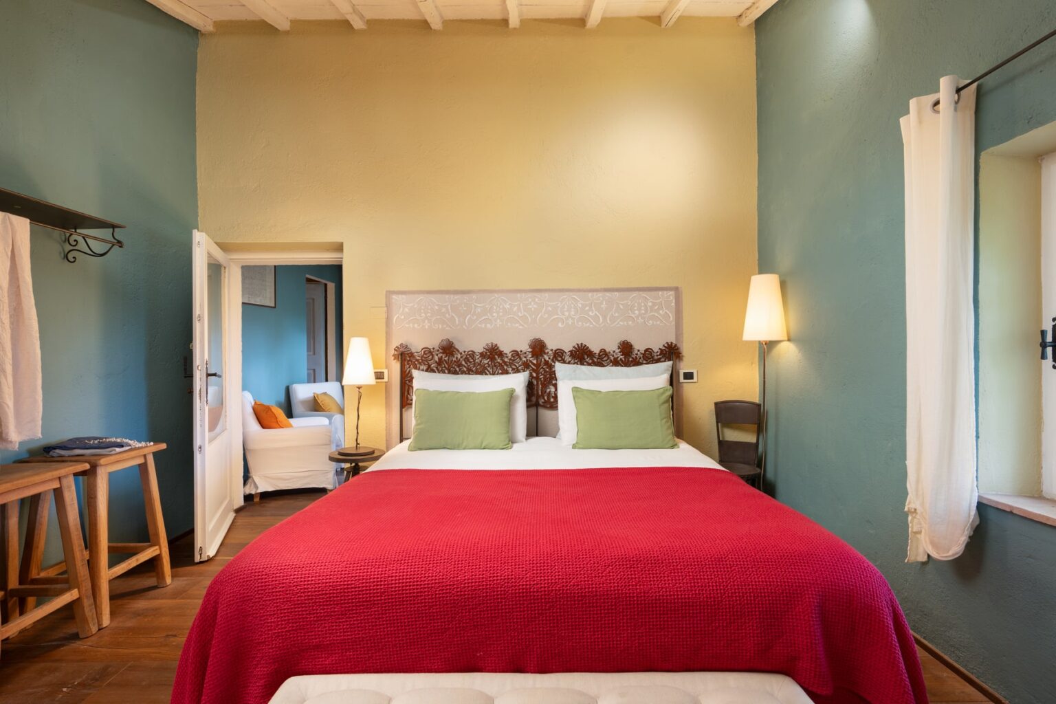 Cozy hotel room featuring a red bedspread, green pillows, and stylish decor, with natural light from windows and a comfortable seating area. Ideal for a relaxing getaway. | Only on SopranoVillas