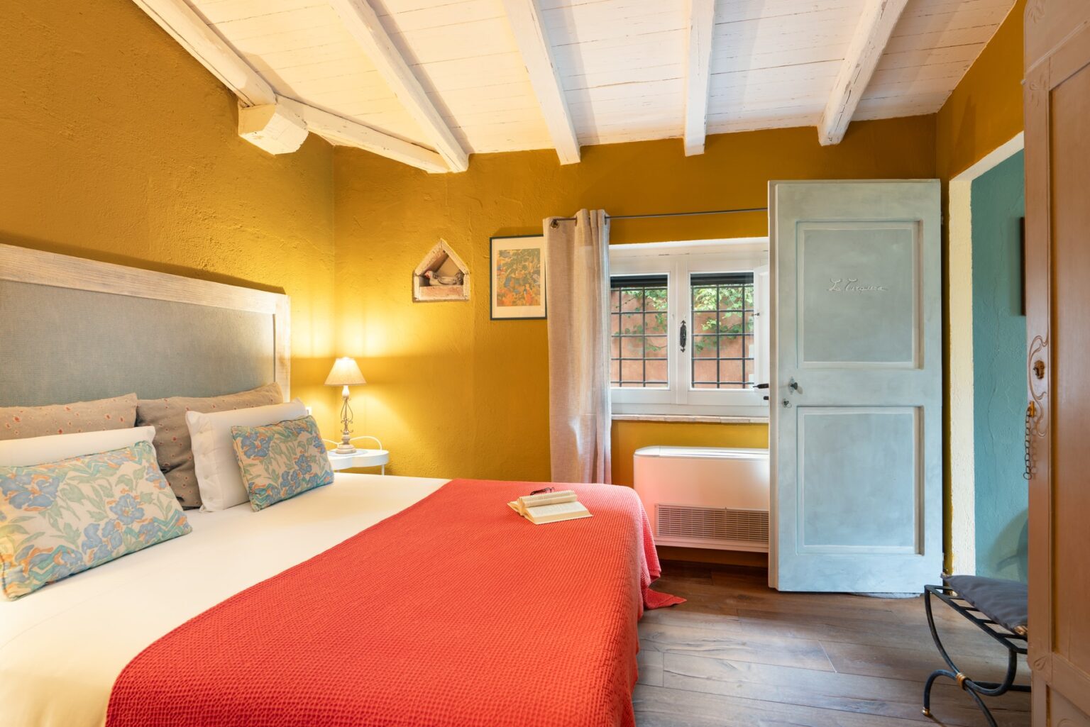Cozy bedroom featuring a king-sized bed with a red throw, warm yellow walls, wooden beams, and a charming window with green shutters, creating a welcoming atmosphere. | Only on SopranoVillas