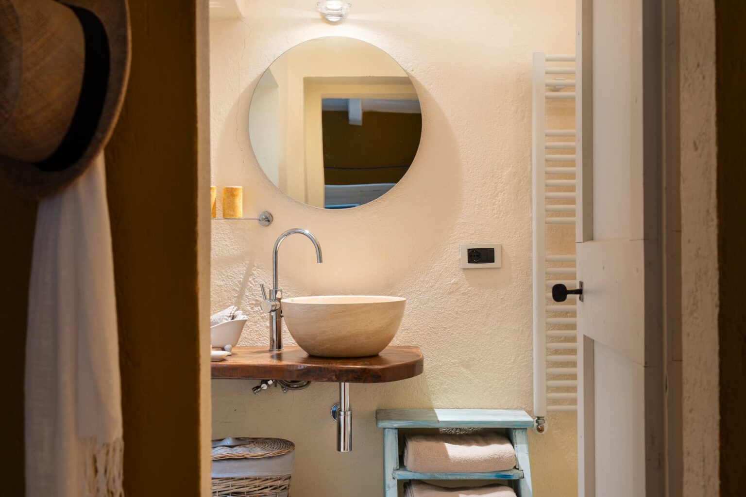 Cozy bathroom featuring a round mirror, a natural stone sink on a wooden countertop, and decorative candles. The space includes a towel rack, a small blue shelf with neatly folded towels, and a woven basket for storage, all set against warm, textured walls. | Only on SopranoVillas