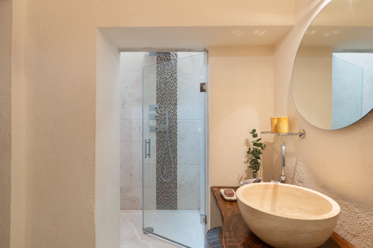 Modern bathroom featuring a spacious glass shower with mosaic tile accents, a round mirror above a stone sink, and warm lighting. | Only on SopranoVillas