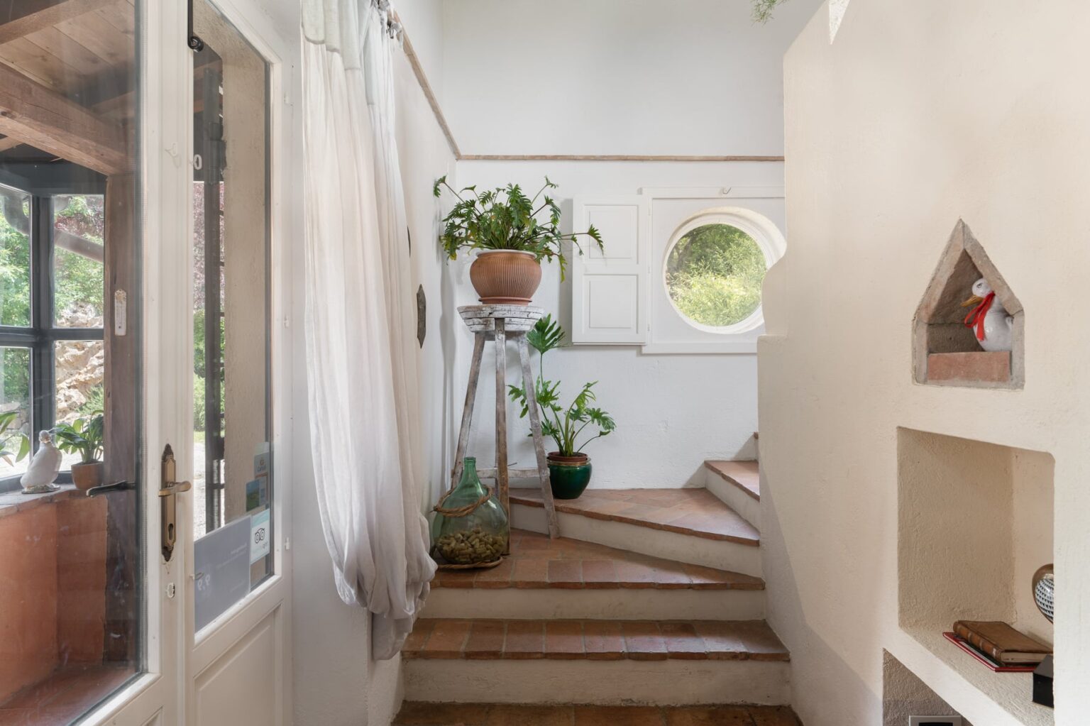Bright and airy interior staircase featuring terracotta tiles, decorative plants in terracotta pots, and a unique round window. The space is illuminated by natural light, creating a welcoming atmosphere. A cozy nook displays decorative items, enhancing the home's charm. | Only on SopranoVillas