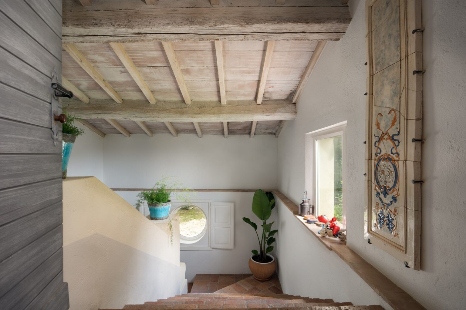 Interior view of a stairwell featuring rustic wooden beams, a round window, and potted plants, creating a warm and inviting atmosphere. | Only on SopranoVillas