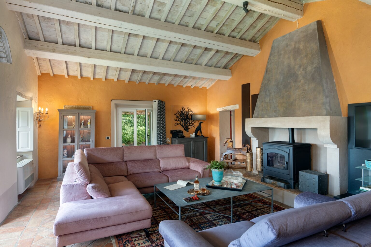 Spacious and inviting living room featuring a pink sectional sofa, rustic wooden ceiling, and a stone fireplace. The warm orange walls create a cozy atmosphere, complemented by natural light from large windows and stylish decor elements. | Only on SopranoVillas