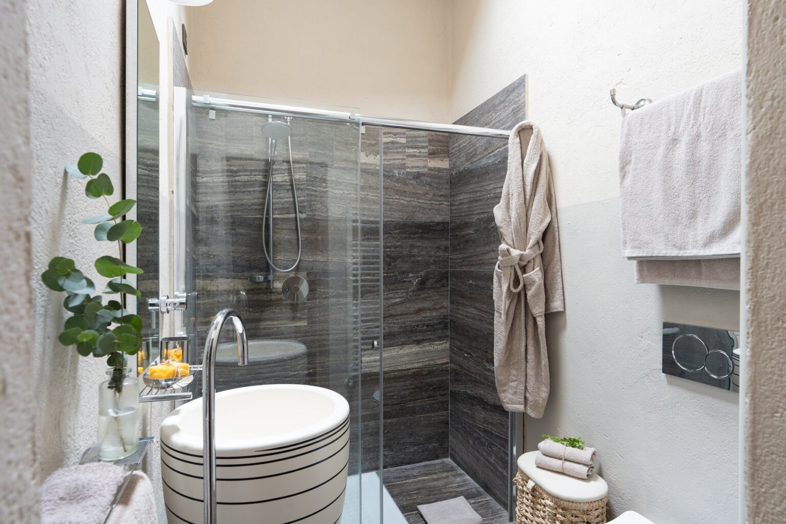 Modern bathroom featuring a glass shower enclosure, a round sink with striped design, plush towels, and a robe hanging on the wall. Greenery adds a touch of nature, enhancing the contemporary aesthetic. Ideal for home renovation inspiration. | Only on SopranoVillas